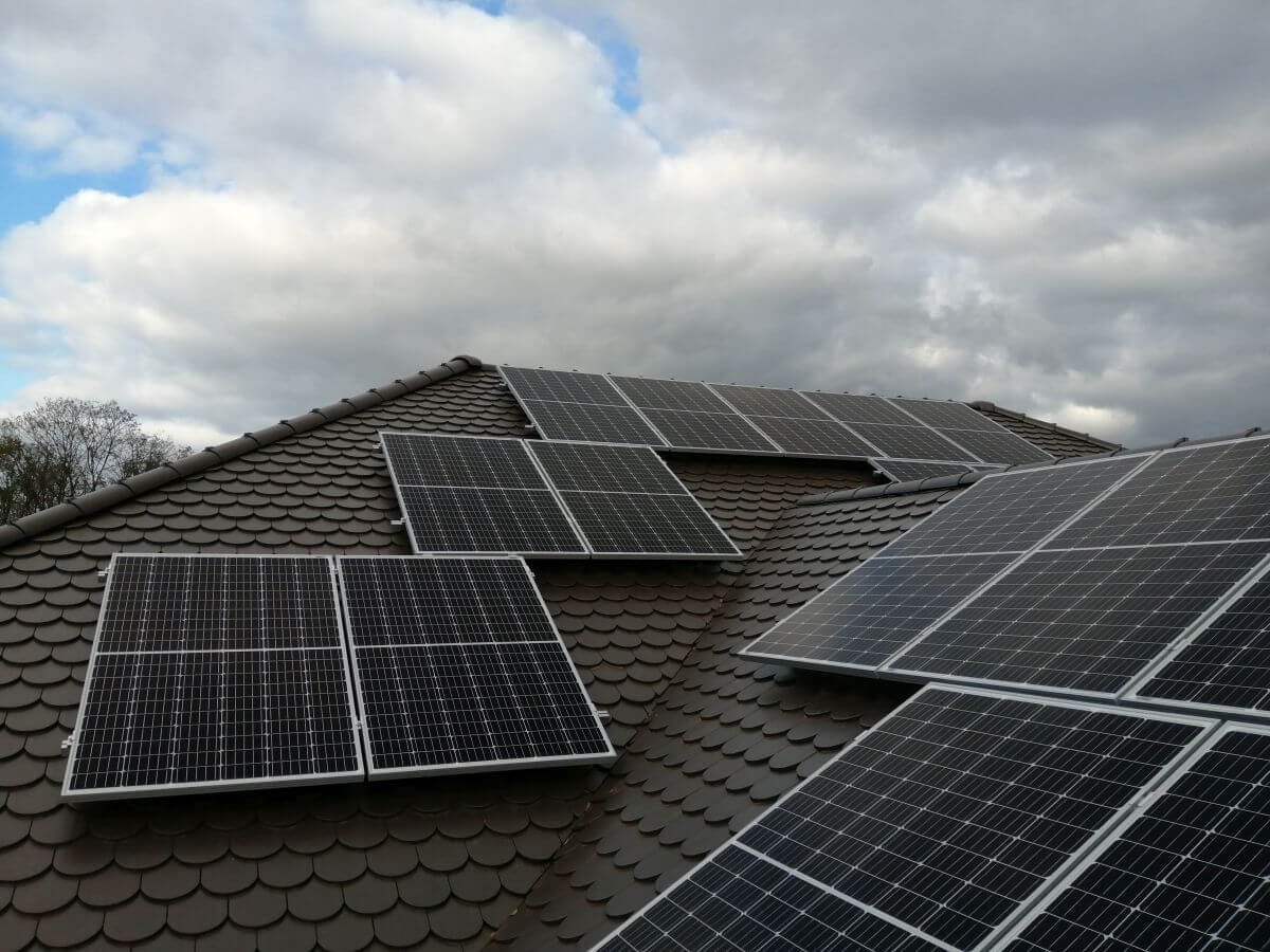 Panele słoneczne zamontowane na brązowym dachu z dachówki ceramicznej, widok z dołu na tle pochmurnego nieba.