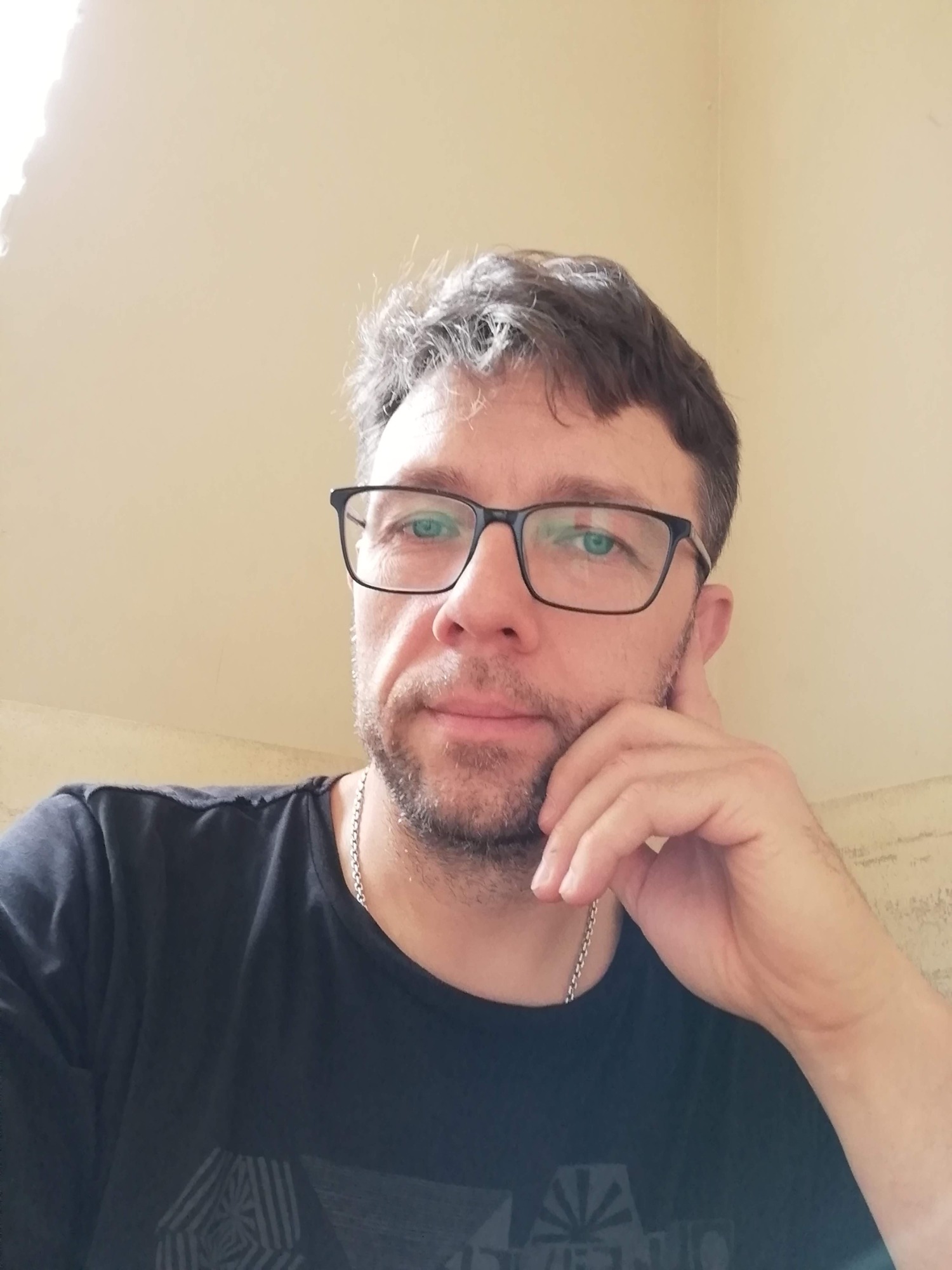 Portret mężczyzny w okularach, z lekkim zarostem i srebrnym łańcuszkiem na szyi, trzymającego dłoń przy policzku, spoglądającego w obiektyw.