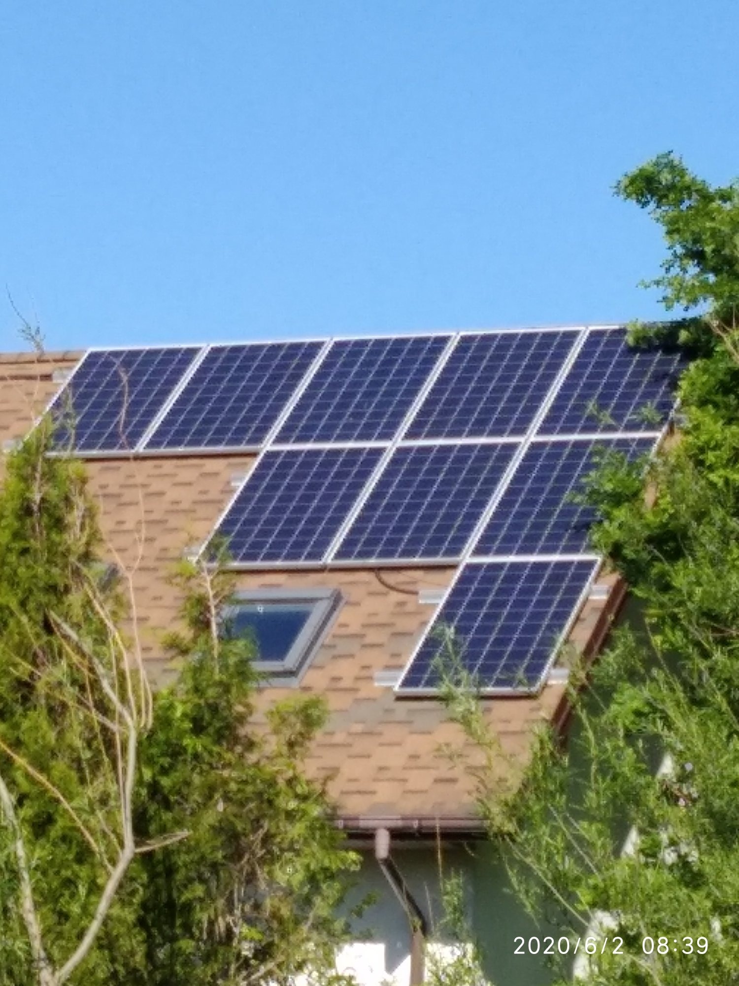 Panele słoneczne na brązowym dachu domu jednorodzinnego z oknem dachowym, częściowo zasłonięte przez drzewa, w słoneczny dzień.