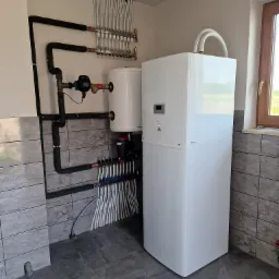 Biała pompa ciepła typu monoblok z podłączonymi rurami grzewczymi i rozdzielaczem, zamontowana w pomieszczeniu z szarą glazurą, widok na okno.