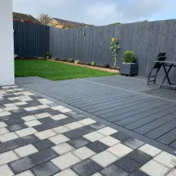 Nowoczesny taras z desek kompozytowych w odcieniach szarości, przylegający do trawnika i ogrodzenia pomalowanego na ciemny kolor, z pojedynczym słonecznikiem przy płocie.