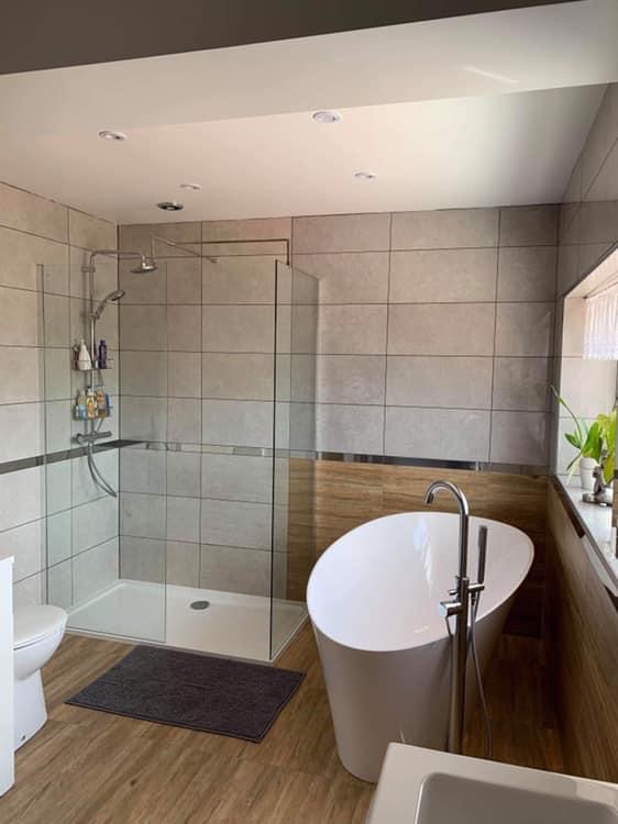 Nowoczesna łazienka z wolnostojącą białą wanną, kabiną prysznicową z szklaną ścianką, toaletą i drewnopodobnymi panelami na podłodze oraz części ściany, oświetlona punktowymi lampami sufitowymi.