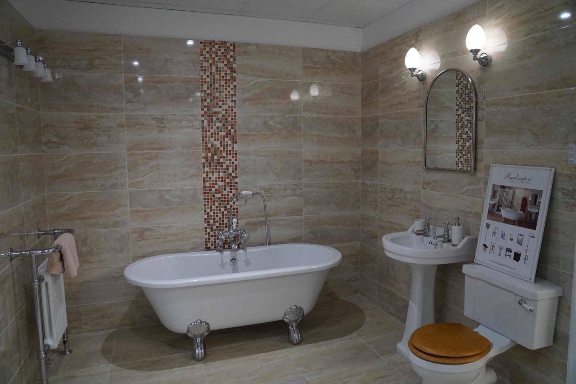 Klasyczna łazienka z wanną na lwich nóżkach, umywalką na postumencie i toaletą z drewnianą deską, wykończona beżowymi płytkami z mozaikowym pasem, oświetlona chromowanymi lampami.