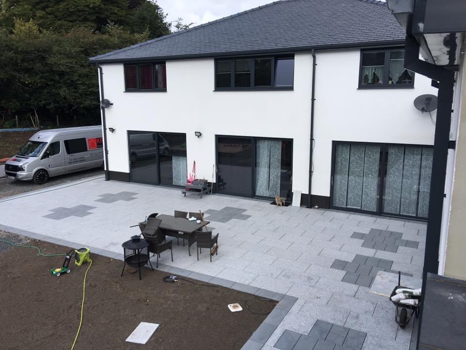 Nowoczesny dom z białymi ścianami i grafitową stolarką okienną, widok na taras z szarej kostki z wzorem krzyży, meble ogrodowe, trawnik z kosiarką i zaparkowany bus firmy w tle.