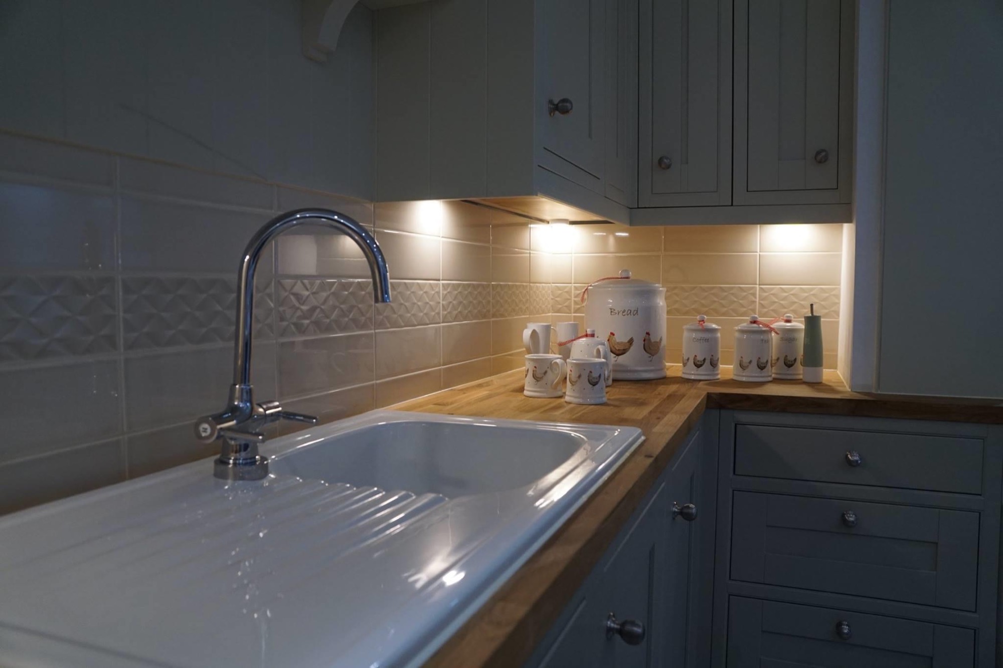 Biały, ceramiczny zlew z chromowaną baterią w jasnej kuchni z drewnianym blatem, podświetlonymi płytkami i pojemnikami na przyprawy z motywem kur