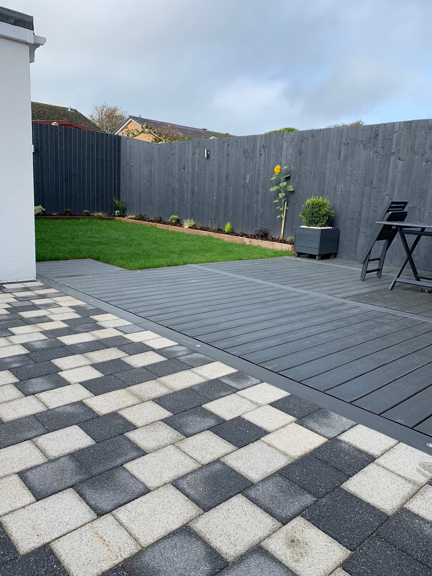 Nowoczesny taras z desek kompozytowych w odcieniach szarości, przylegający do trawnika i ogrodzenia pomalowanego na ciemny kolor, z pojedynczym słonecznikiem przy płocie.