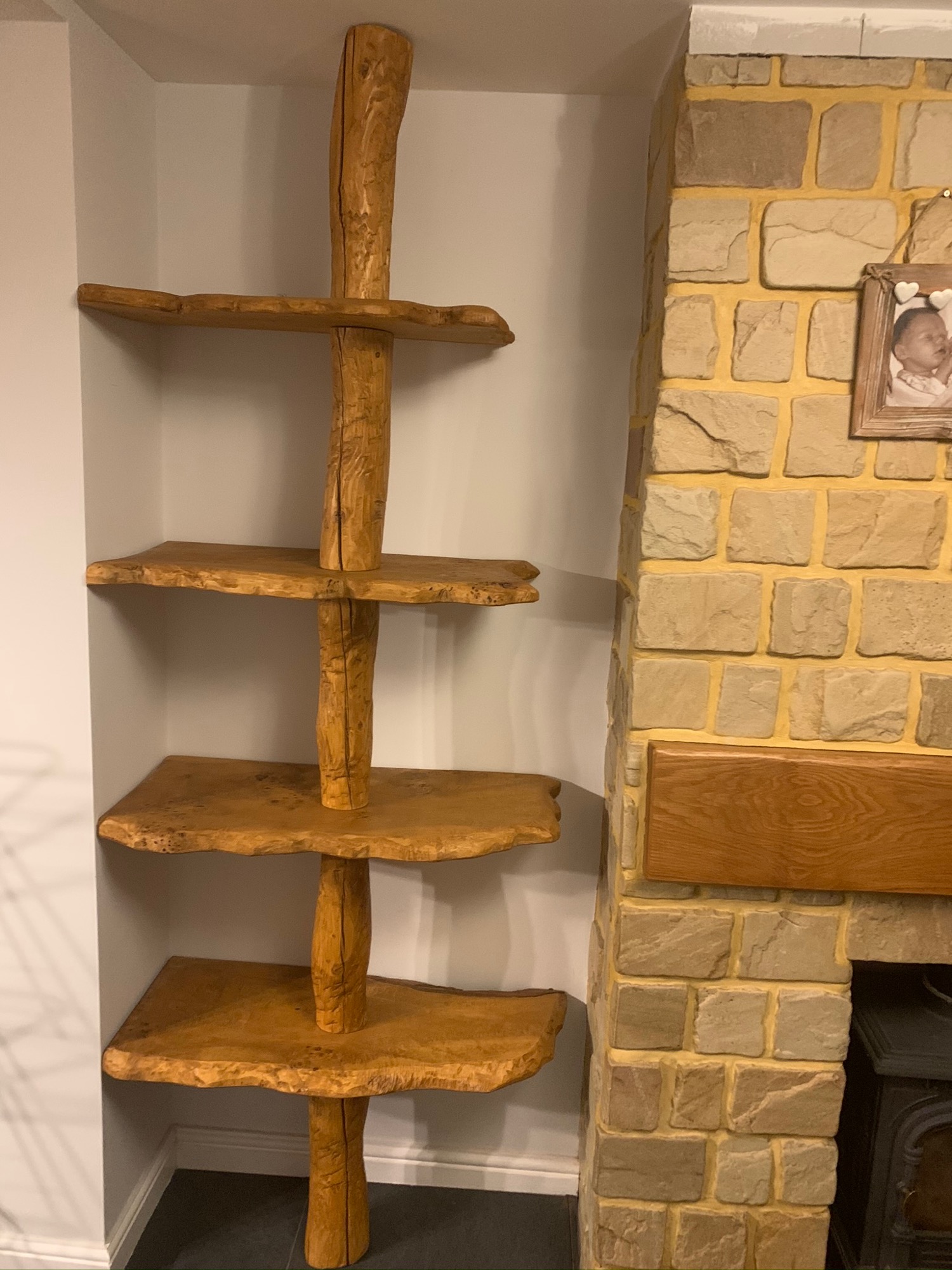 Ręcznie robiony regał z drewna o nieregularnych kształtach, wkomponowany w narożnik pokoju obok kamiennej ściany kominka.