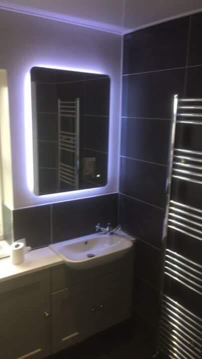 Nowoczesna łazienka z ciemnymi płytkami, podświetlanym lustrem i grzejnikiem drabinkowym, widok na umywalkę z szafką.