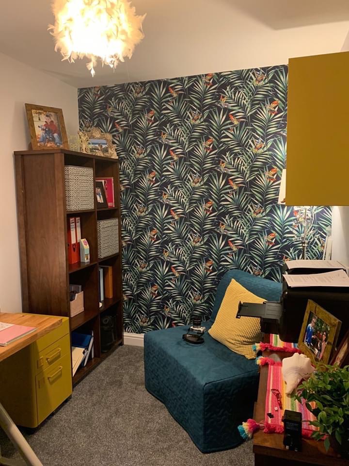 Pokój z ciemną tapetą w tropikalne liście, drewnianym regałem, żółtym biurkiem, turkusowym siedziskiem i puszystą lampą sufitową.