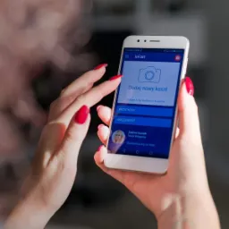 Kobieta trzyma biały smartfon z aplikacją inFakt wyświetlającą opcje dodawania kosztów, przychodów i księgowości, z czerwonym manicurem na paznokciach.