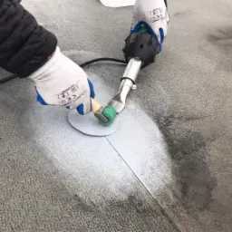 Zgrzewanie membrany dachowej za pomocą zgrzewarki ręcznej i dociskania rolką; widoczne rękawice ochronne i kabel zasilający.