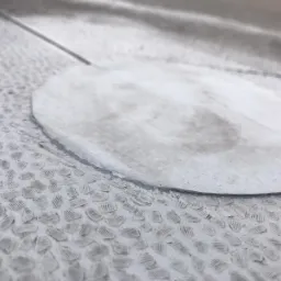 Szczegółowe ujęcie okrągłej, białej membrany izolacyjnej nałożonej na szorstką, teksturowaną powierzchnię, prawdopodobnie fundamentu budynku, z widocznymi śladami użytkowania i delikatnymi...