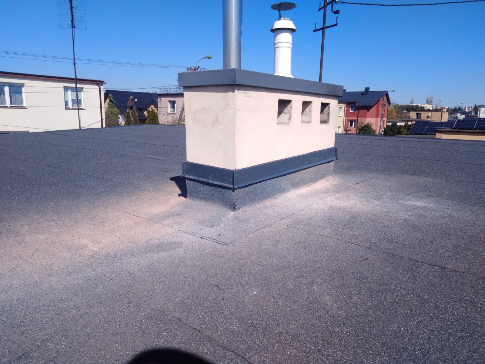 Płaski dach pokryty papą z kominem wentylacyjnym i wywietrznikami, widoczne ślady posypki bitumicznej.