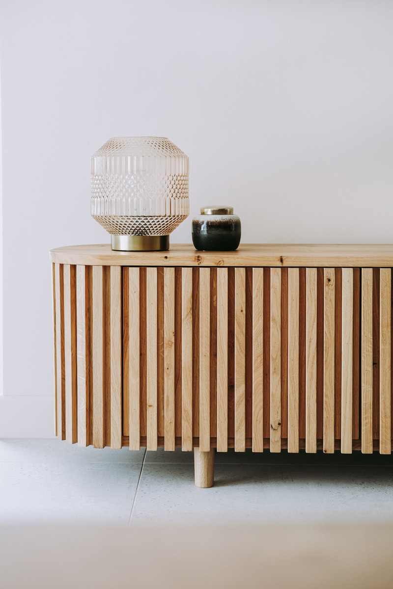 Drewniana komoda z lamelami na okrągłych nóżkach, na blacie szklana lampa w geometryczne wzory i ceramiczny pojemnik z metalową pokrywką.
