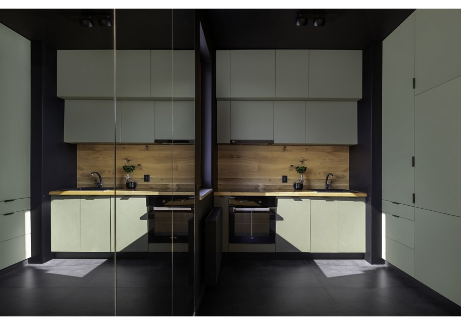 Nowoczesna kuchnia w odcieniach zieleni i czerni, z drewnianym panelem nad blatem i minimalistycznym wystrojem, widziana z perspektywy odbicia w szklanej powierzchni.