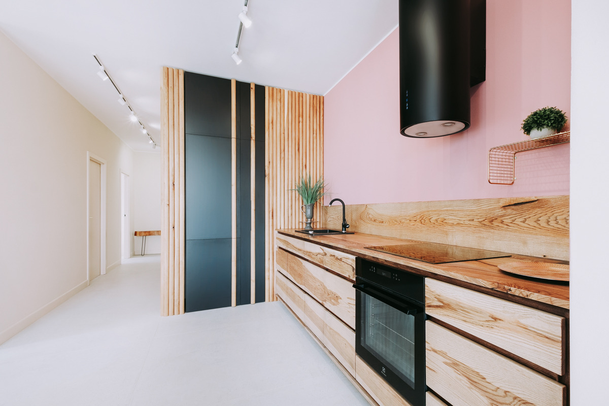 Nowoczesna kuchnia z frontami z jasnego drewna o wyraźnym usłojeniu, czarny piekarnik w zabudowie, różowa ściana i czarny okrągły okap, na blacie czarna płyta indukcyjna i metalowa półka z rośliną,...