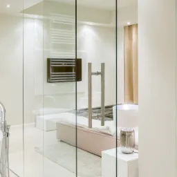 Nowoczesna łazienka z wolnostojącą wanną, chromowaną armaturą i grzejnikiem drabinkowym na beżowej ścianie, widok na sypialnię przez szklaną ścianę i białą szafę wnękową.