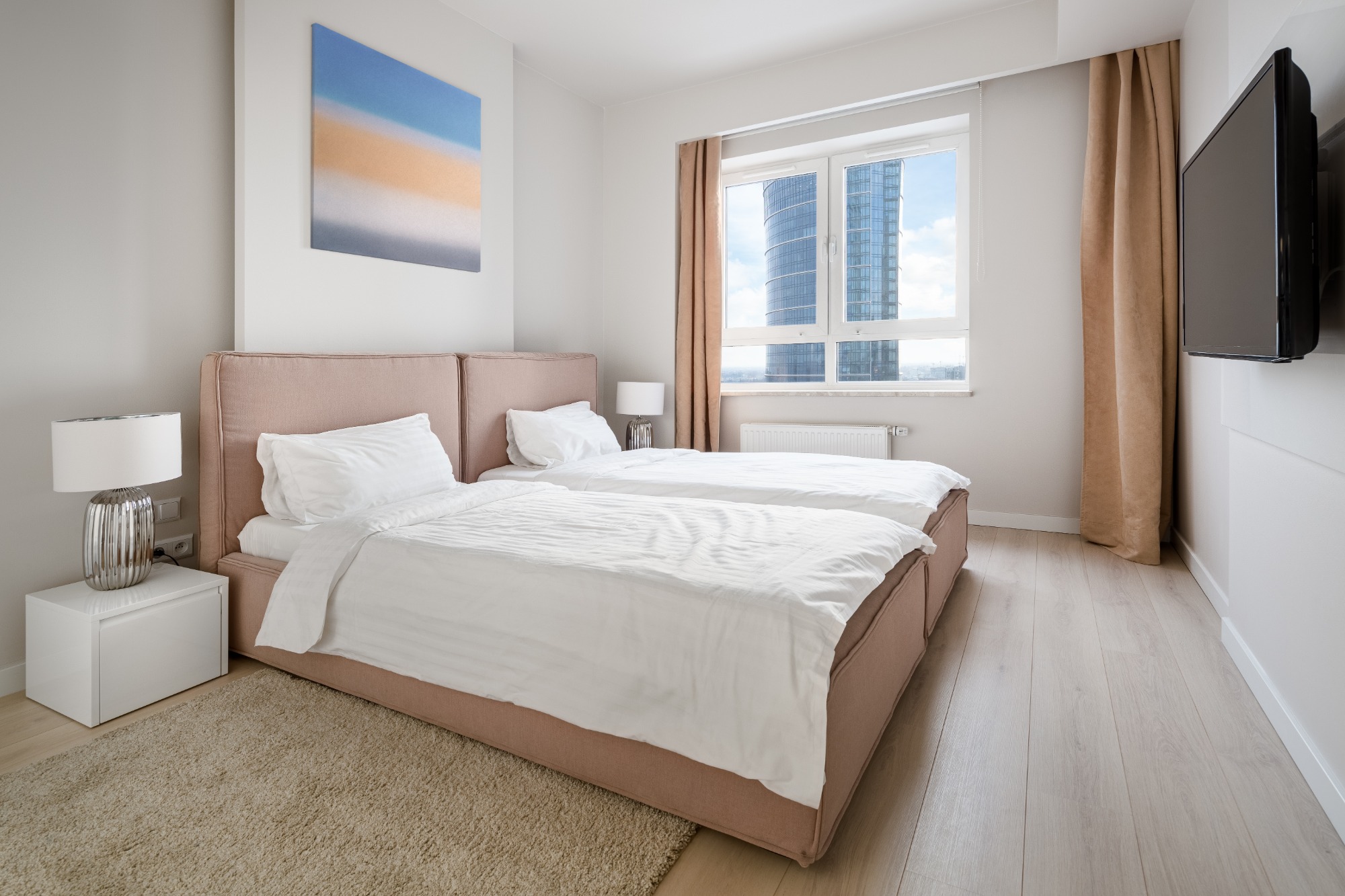 Jasna sypialnia z dwoma pojedynczymi łóżkami z różowymi zagłówkami, obraz na ścianie i widok na wieżowiec za oknem.