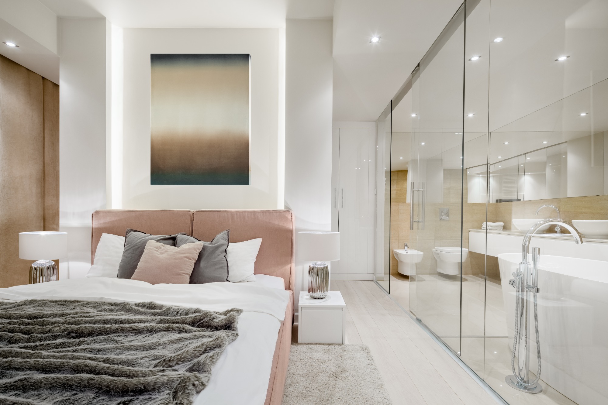 Nowoczesna sypialnia z łazienką oddzieloną szklaną ścianą, łóżko z różowym zagłówkiem, szarą narzutą i obrazem na ścianie, widok na białą armaturę łazienkową.