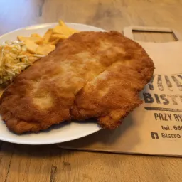 Smażony sznycel na białym talerzu z frytkami i surówką z kapusty, obok papierowa torba z nadrukowanym logo 'Bistro Przy Ryneczku' na drewnianym stole.