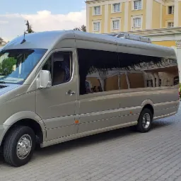 Srebrny Mercedes-Benz Sprinter z przyciemnianymi szybami zaparkowany na kostce brukowej przed żółtym budynkiem z kolumnami i dekoracyjnym oknem.