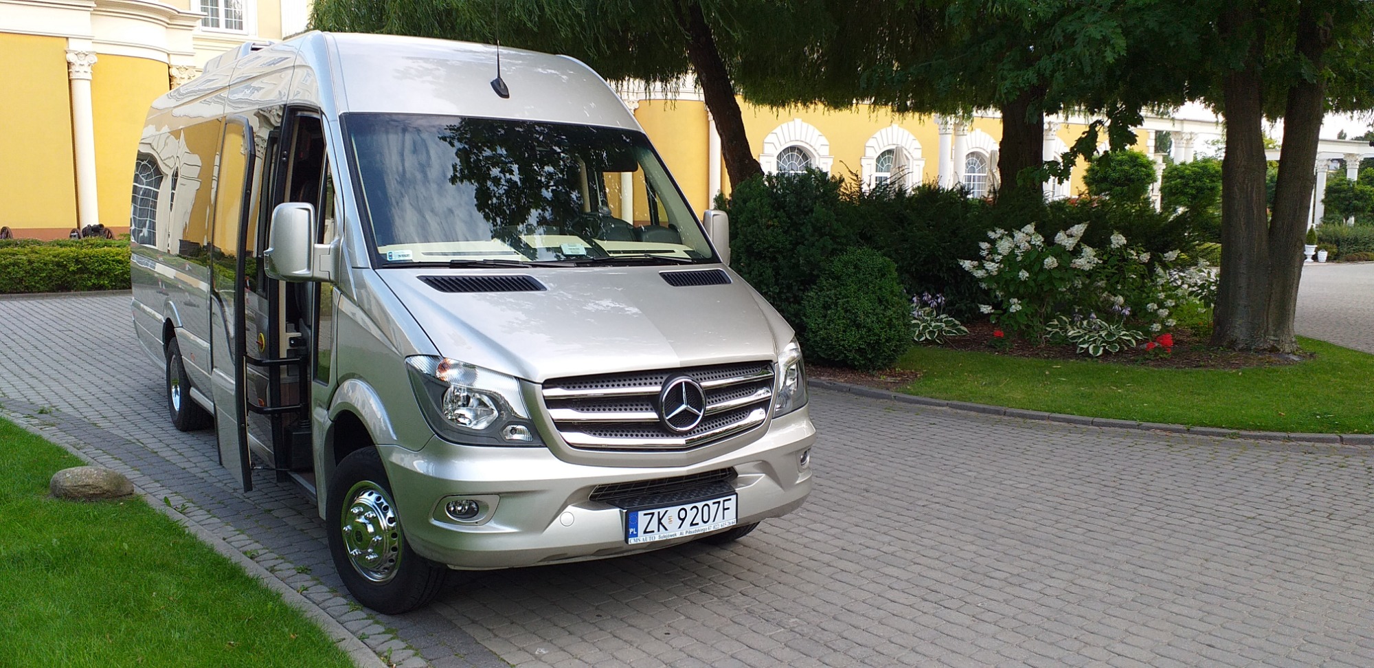 Srebrny bus marki Mercedes z otwartymi drzwiami, zaparkowany na brukowanej drodze przed budynkiem z kolumnami i zielenią.