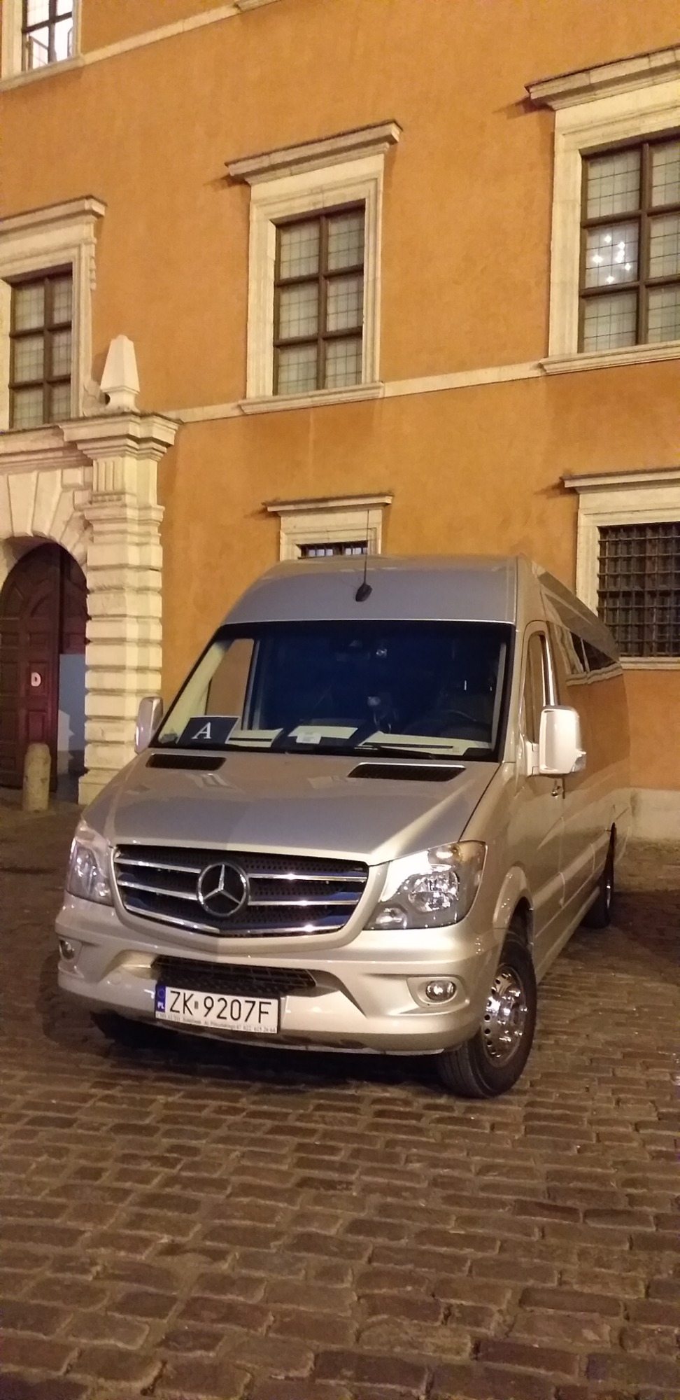Srebrny bus Mercedes-Benz Sprinter zaparkowany na brukowanej ulicy przed pomarańczową kamienicą z oknami w białych ramach.