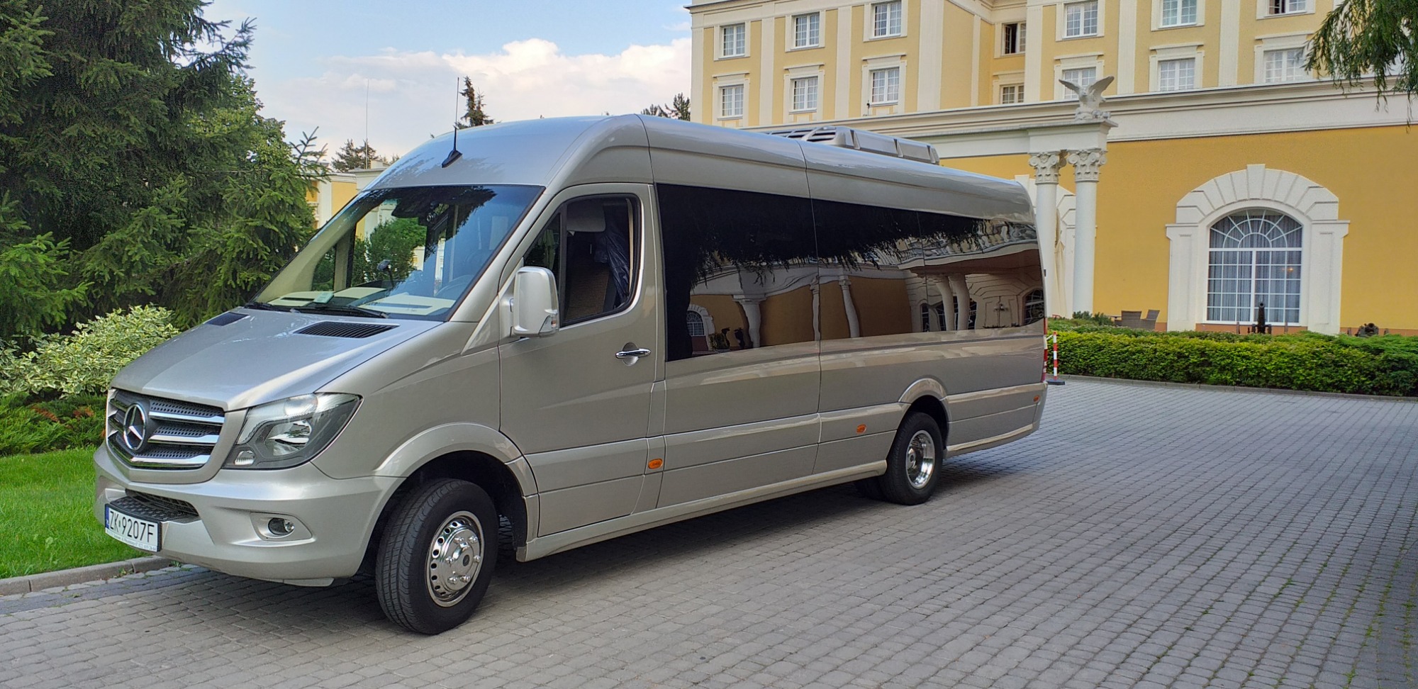 Srebrny Mercedes-Benz Sprinter z przyciemnianymi szybami zaparkowany na kostce brukowej przed żółtym budynkiem z kolumnami i dekoracyjnym oknem.