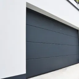 Zamknięta, grafitowa brama garażowa segmentowa z poziomymi przetłoczeniami, wkomponowana w nowoczesną, białą bryłę budynku z ciemnymi akcentami.