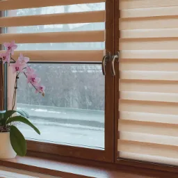 Brązowe okno z roletami dzień i noc w beżowych odcieniach, na parapecie stoi biała doniczka z kwitnącą orchideą.