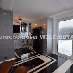 Wnętrze domu z czarno-białą marmurową podłogą w geometryczne wzory, widok na kuchnię z wyspą i barowymi stołkami, oraz okno z widokiem na zaśnieżony krajobraz.