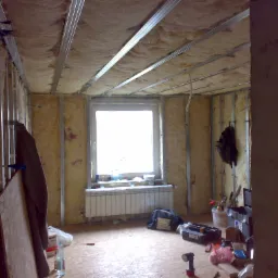 Adaptacja poddasza: widok na wnętrze w trakcie ocieplania wełną mineralną, widoczne metalowe profile konstrukcyjne sufitu i ścian, okno z grzejnikiem, narzędzia i materiały budowlane na podłodze.