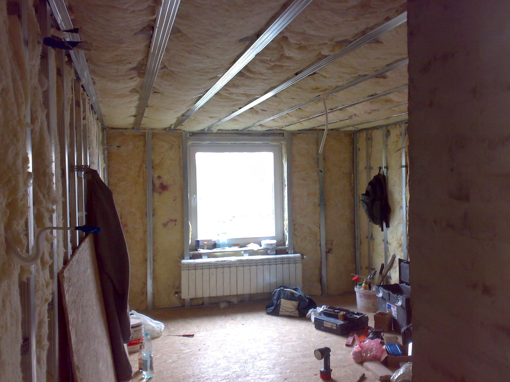Adaptacja poddasza: widok na wnętrze w trakcie ocieplania wełną mineralną, widoczne metalowe profile konstrukcyjne sufitu i ścian, okno z grzejnikiem, narzędzia i materiały budowlane na podłodze.