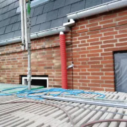 Ceglana ściana budynku z zamontowaną rynną i czerwoną rurą spustową, widoczne rusztowanie i okno zabezpieczone folią.