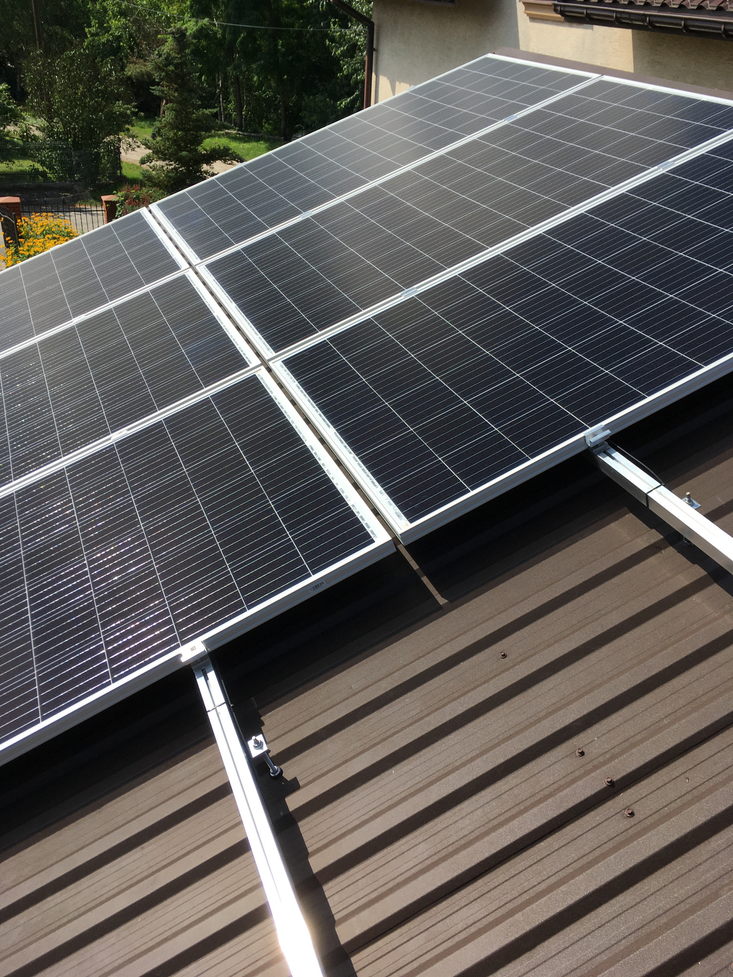 Panele słoneczne zamontowane na brązowym dachu z blachy trapezowej, widoczne mocowania i cień paneli.