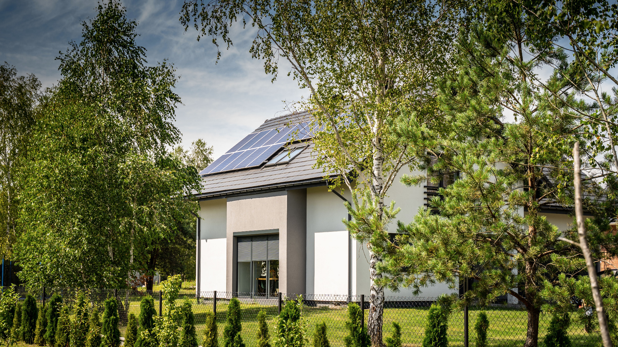 Nowoczesny dom jednorodzinny z panelami słonecznymi na dachu, widoczny zza ogrodzenia i drzew w słoneczny dzień.
