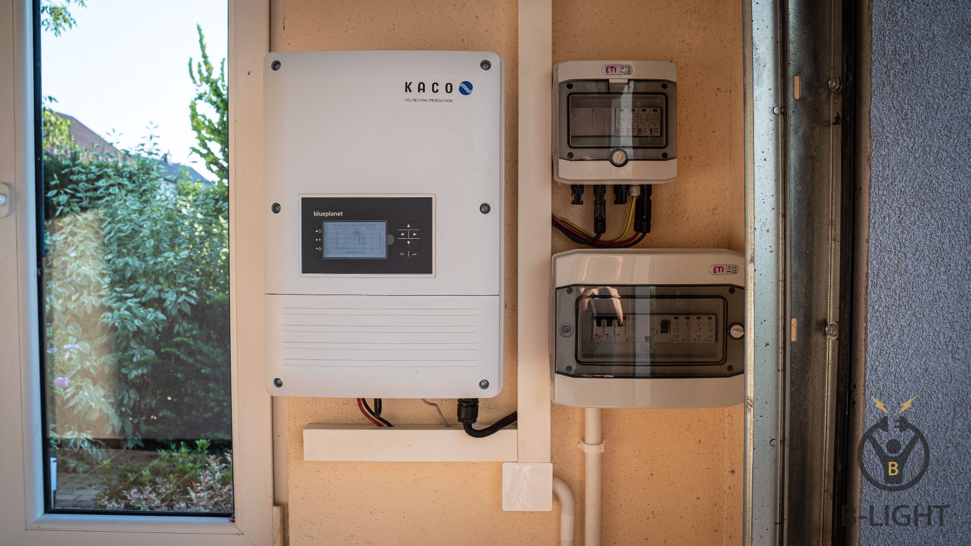 Biały inwerter KACO blueplanet z wyświetlaczem i dwa zabezpieczenia ETI zamontowane na ścianie obok okna z widokiem na zieleń.