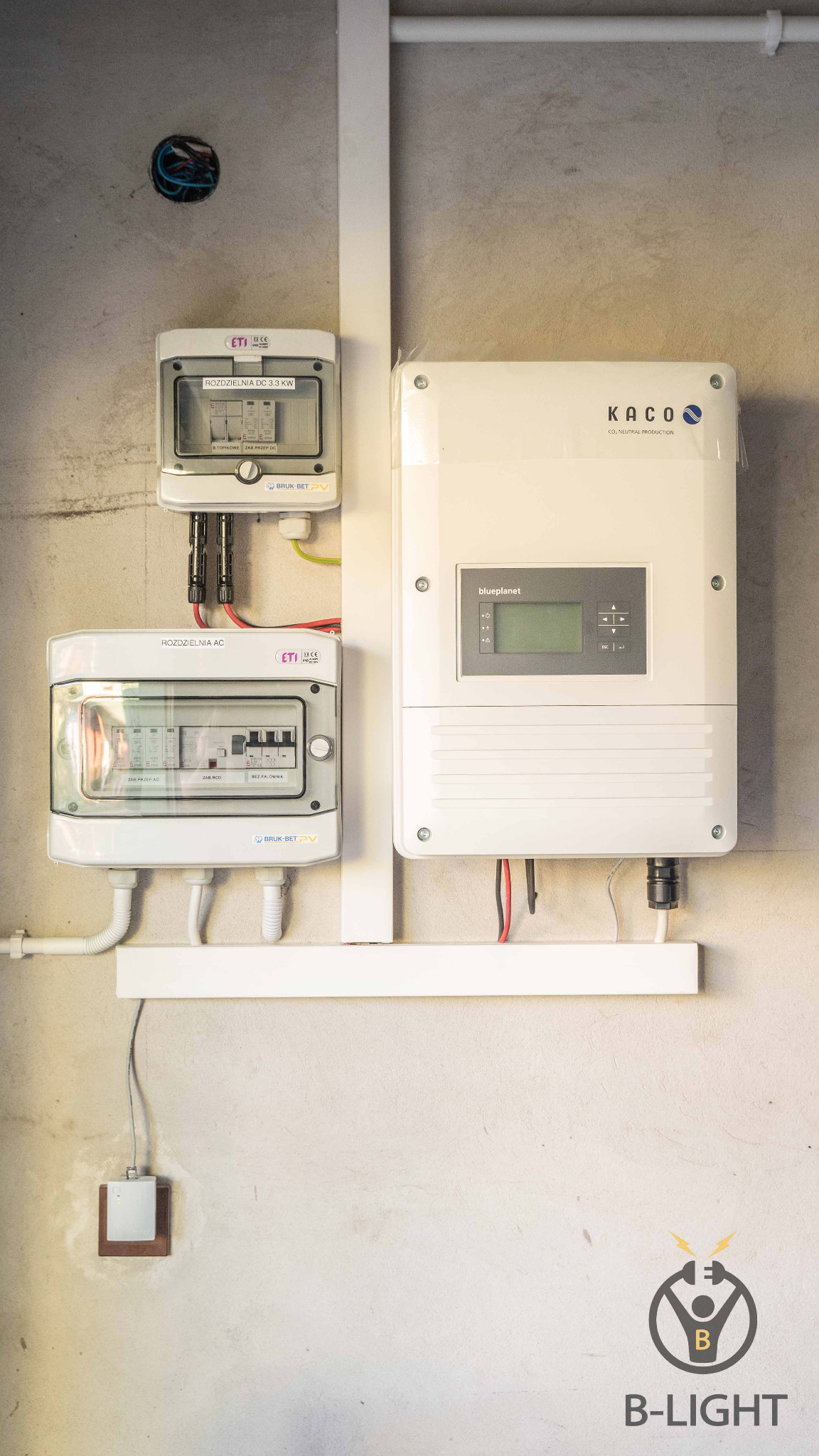 Dwa rozdzielacze AC i DC oraz falownik Kaco Blueplanet zamontowane na ścianie z widocznymi przewodami elektrycznymi i logo firmy B-Light w prawym dolnym rogu.