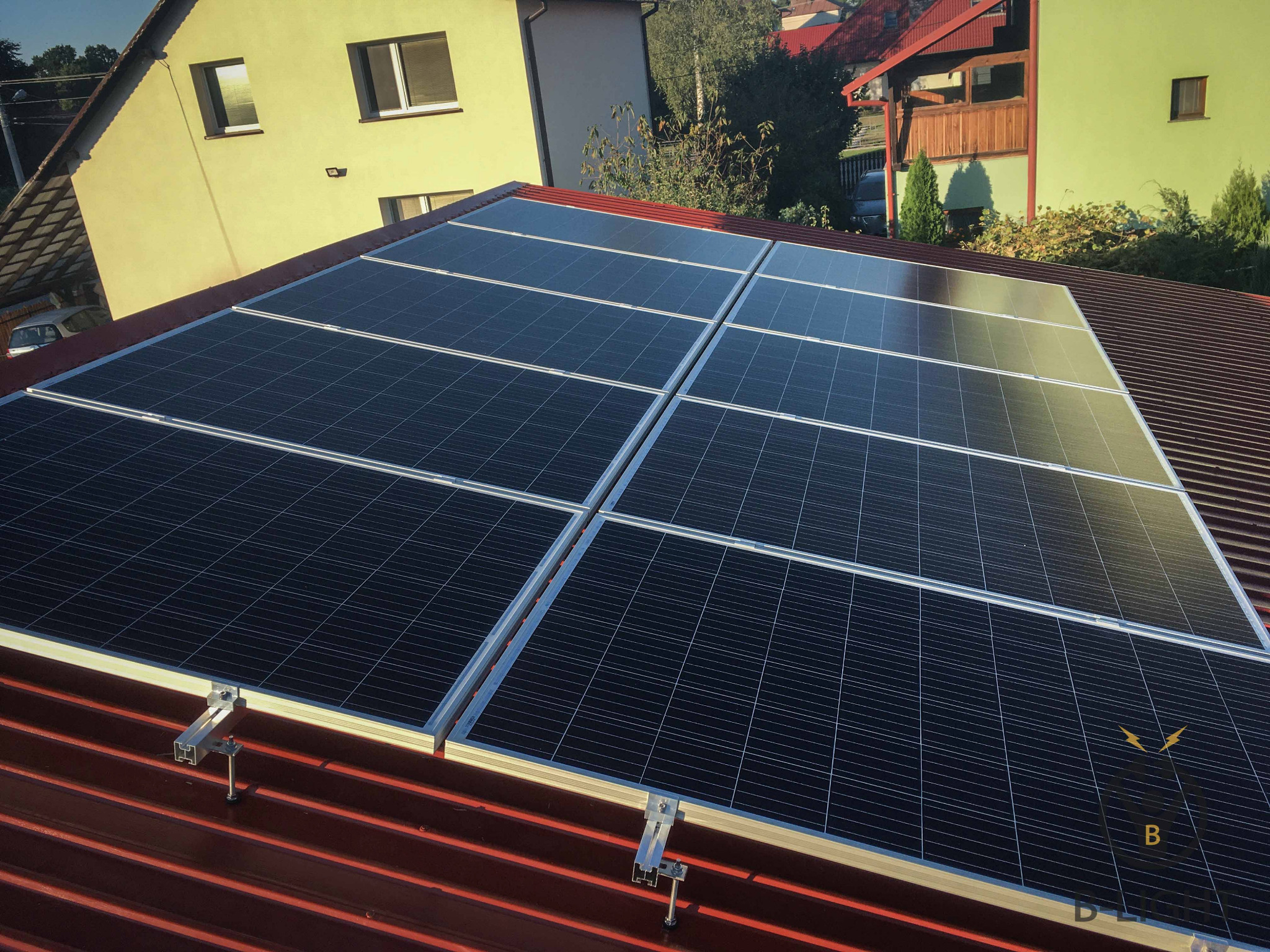 Panele słoneczne zamontowane na czerwonym, blaszany dachu domu jednorodzinnego, widoczne elementy mocujące.