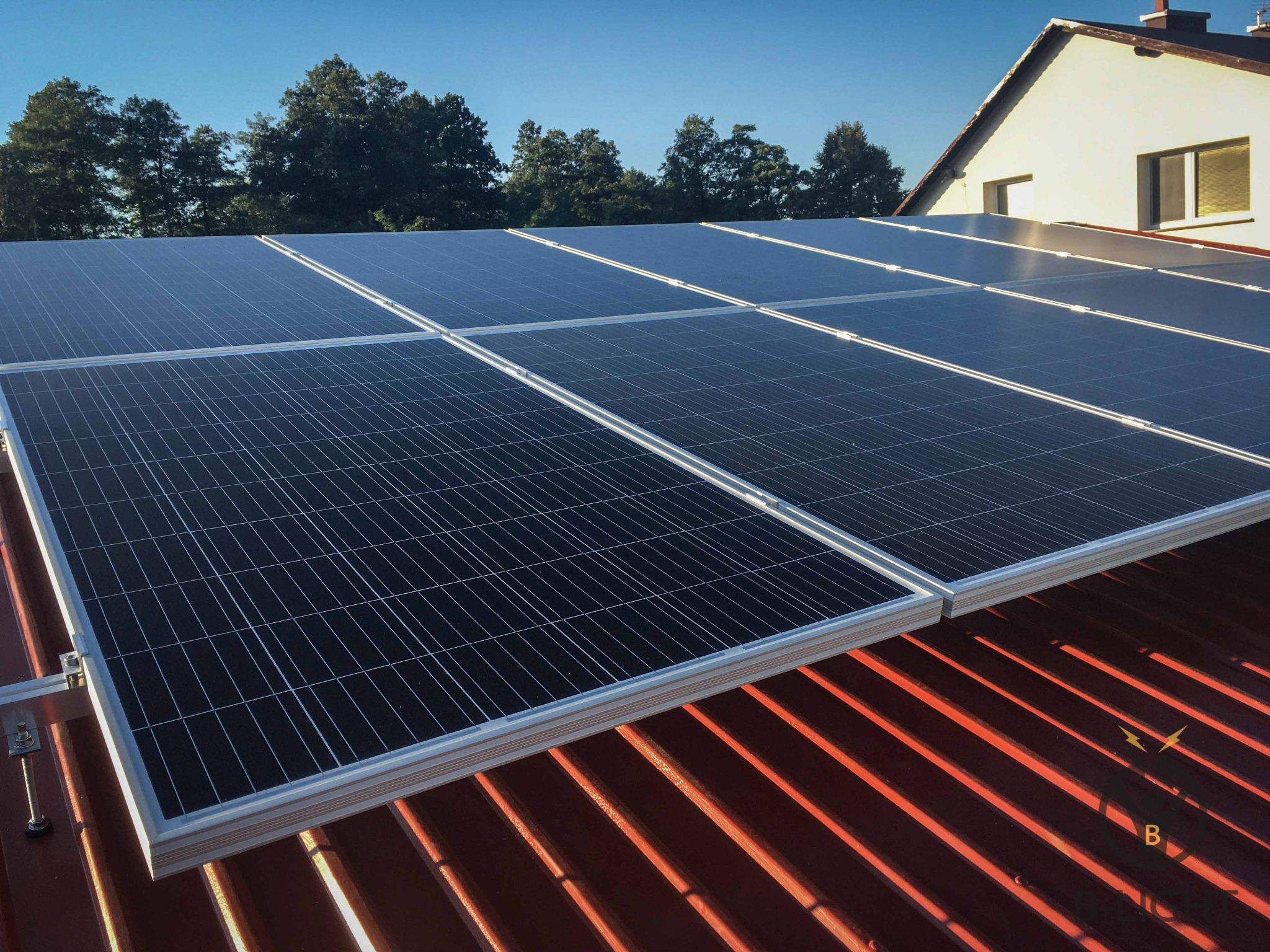 Panele słoneczne na czerwonym dachu blaszanej z widocznym mocowaniem, dom i drzewa w tle, słoneczny dzień.