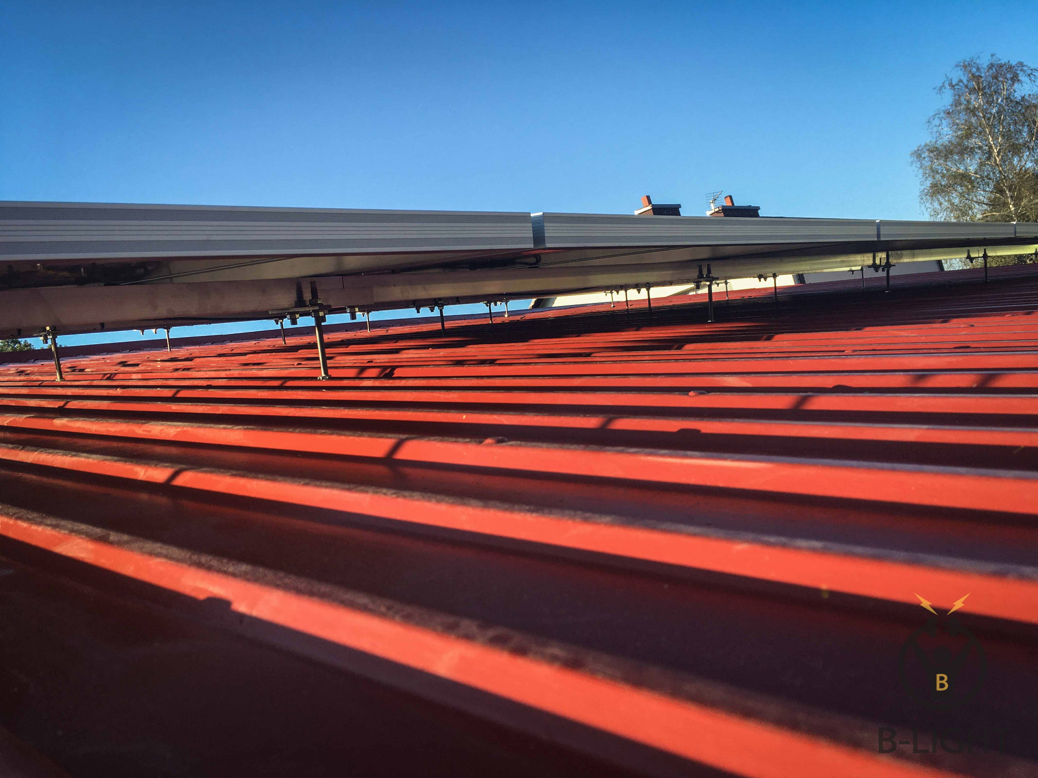 Montaż paneli fotowoltaicznych na czerwonym, metalowym dachu, widoczne mocowania i logo firmy w rogu zdjęcia.