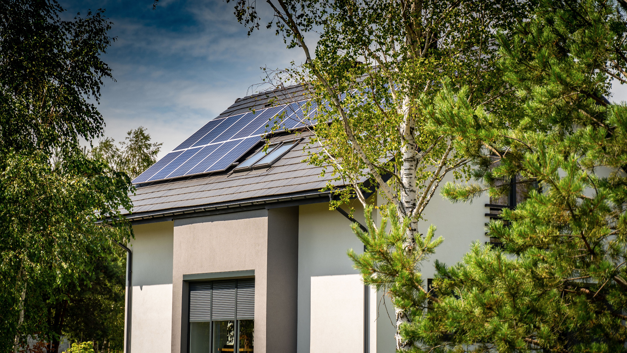Panele słoneczne na dachu nowoczesnego domu jednorodzinnego z oknami dachowymi, widoczne przez korony drzew.