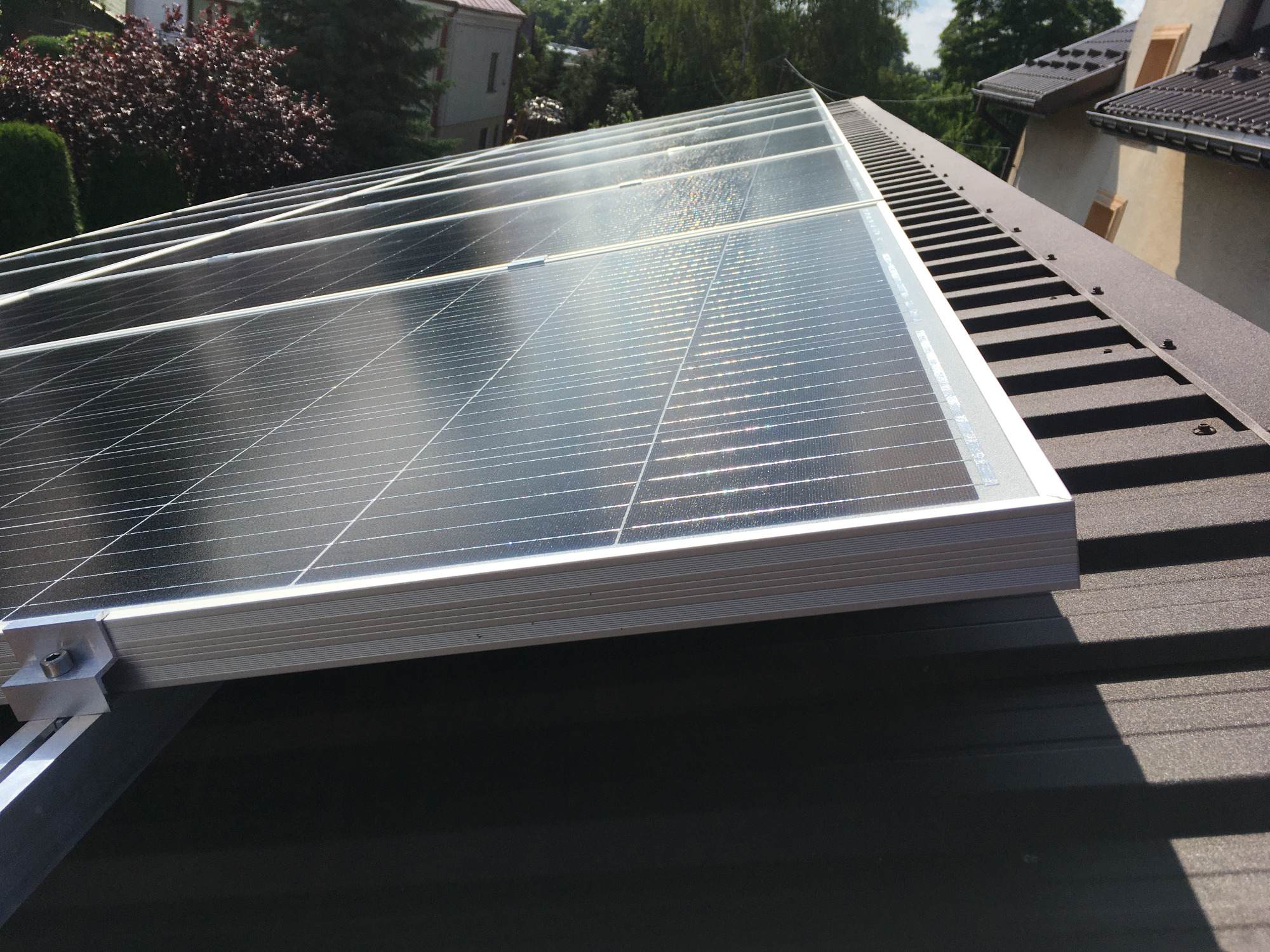 Panele słoneczne zamontowane na brązowym dachu z blachy, widoczne elementy mocujące, budynki w tle.