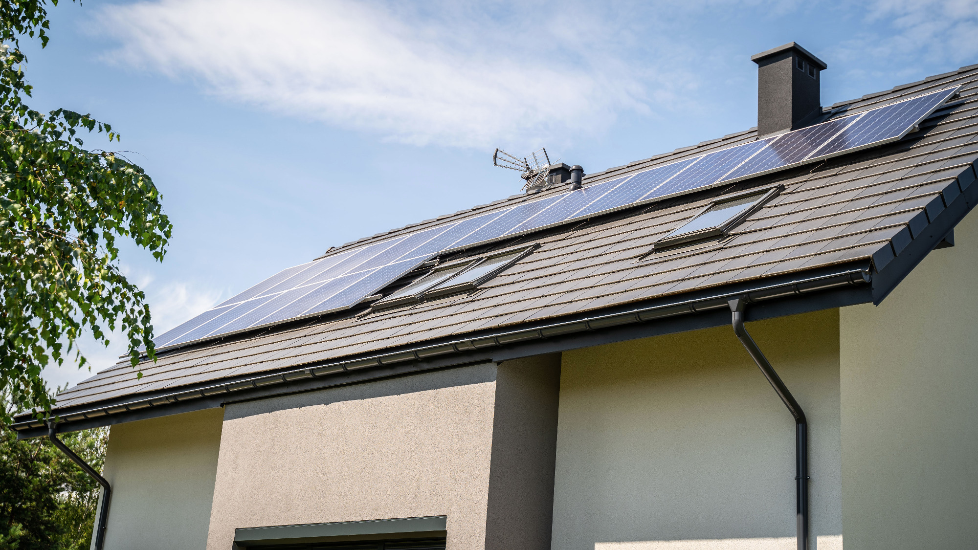 Panele słoneczne na dachu domu jednorodzinnego z brązową dachówką, widoczne okna dachowe, komin i antena telewizyjna.