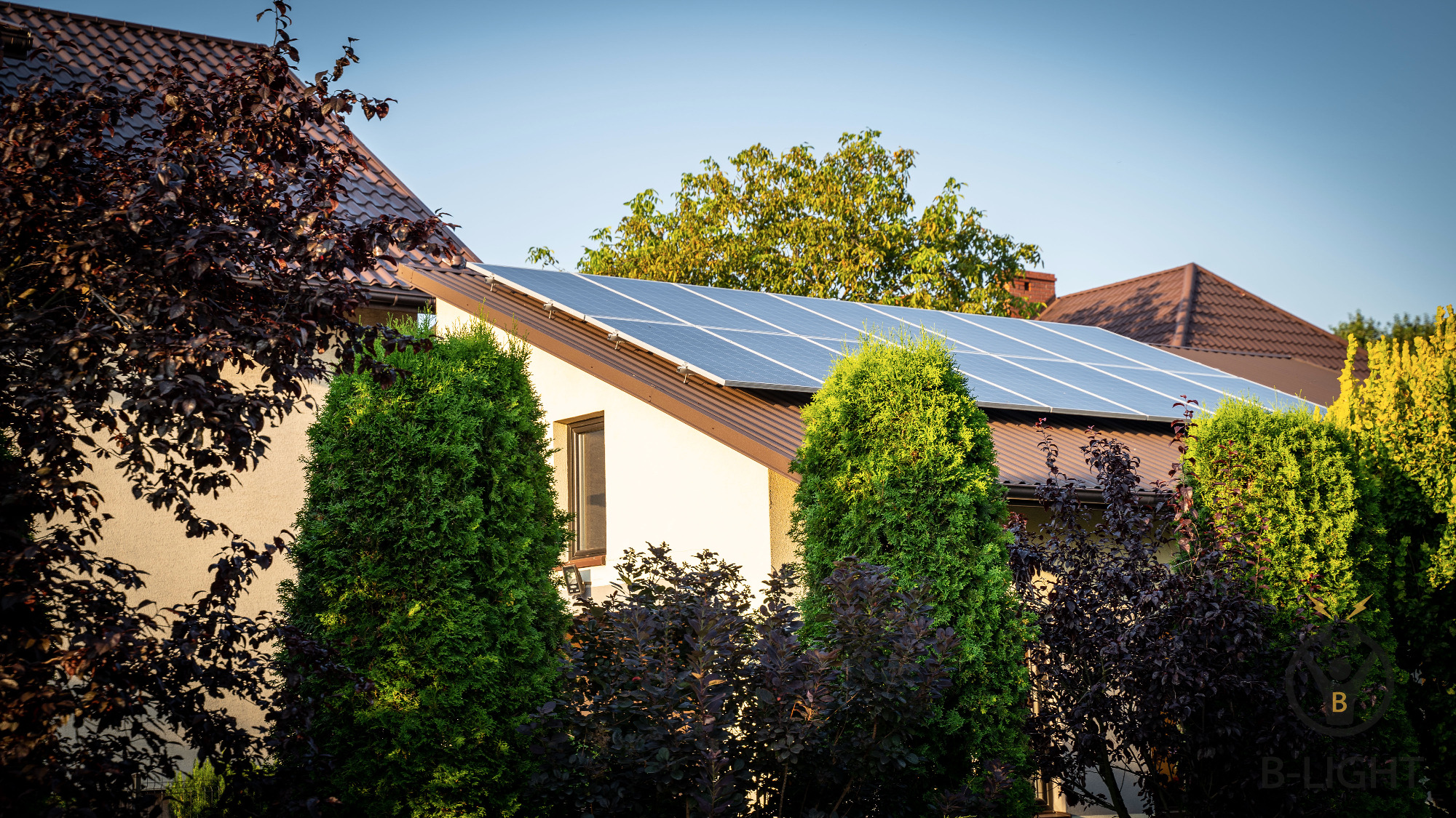 Panele słoneczne na brązowym dachu domu jednorodzinnego, widoczne drzewa iglaste i liściaste w ogrodzie.