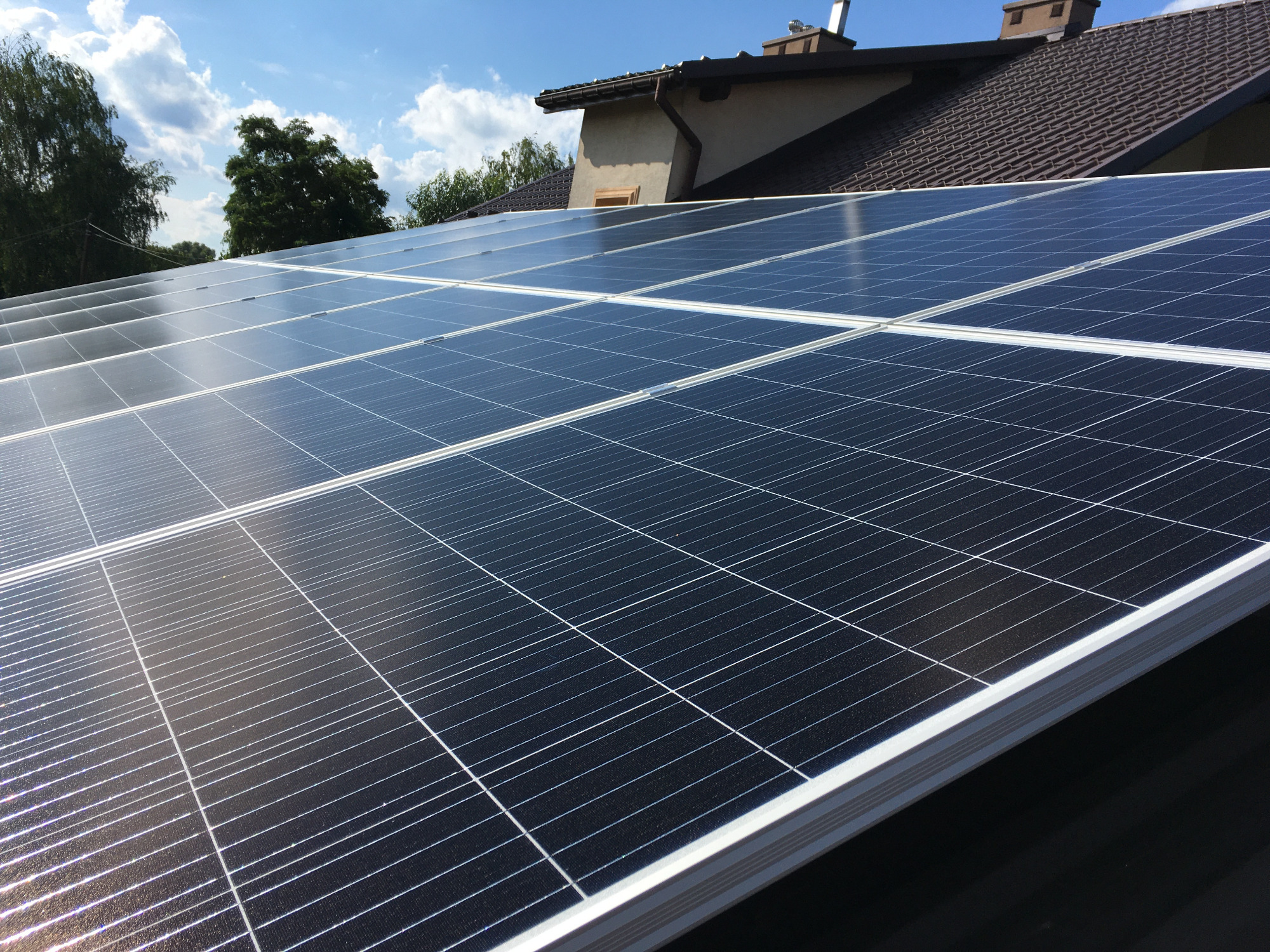 Rząd paneli słonecznych na dachu domu jednorodzinnego w słoneczny dzień, widoczne odbicia nieba na panelach.