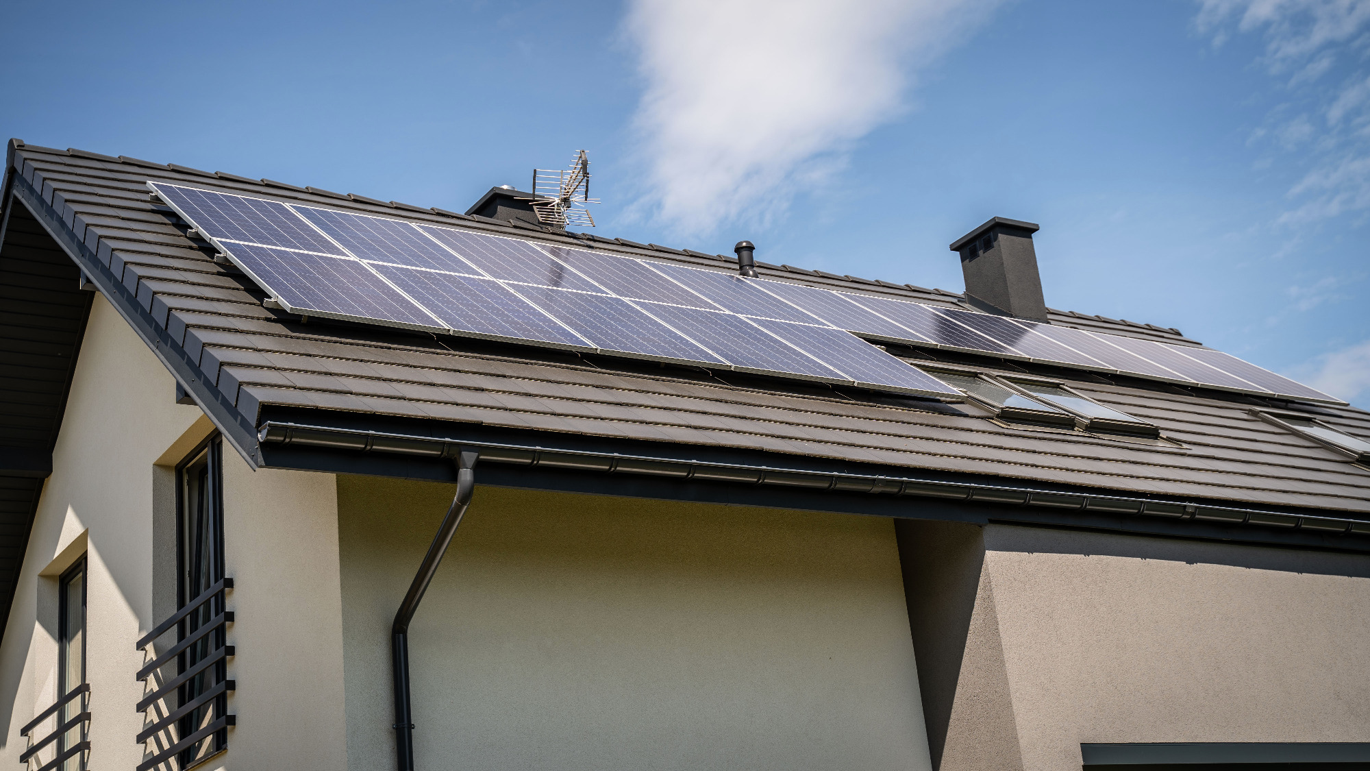 Panele fotowoltaiczne na dachu nowoczesnego domu z brązową dachówką, widoczne okna dachowe i antena telewizyjna.