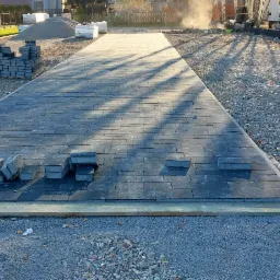 Położona częściowo szara kostka brukowa w jodełkę, z przygotowanym podłożem i stosem kostki obok, w tle budynek mieszkalny i samochód dostawczy.