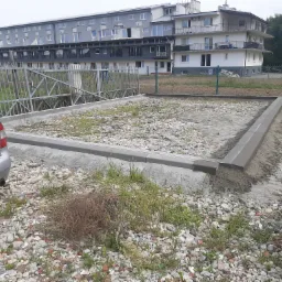 Przygotowanie podłoża pod przyszłą nawierzchnię: obrzeża betonowe osadzone w piasku, zagęszczarka w tle, gruz i roślinność na pierwszym planie, budynek mieszkalny w tle.