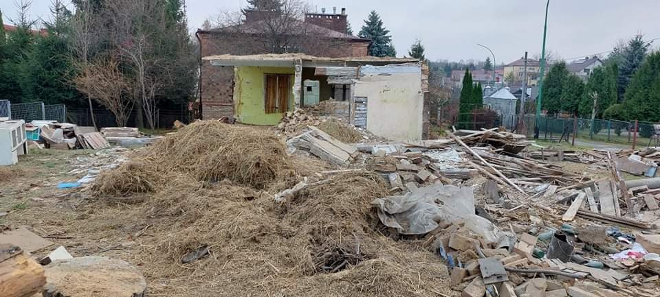 Częściowo zburzony budynek z odsłoniętą zieloną ścianą i stertą gruzu, cegieł i siana na pierwszym planie, w tle zabudowa mieszkalna.